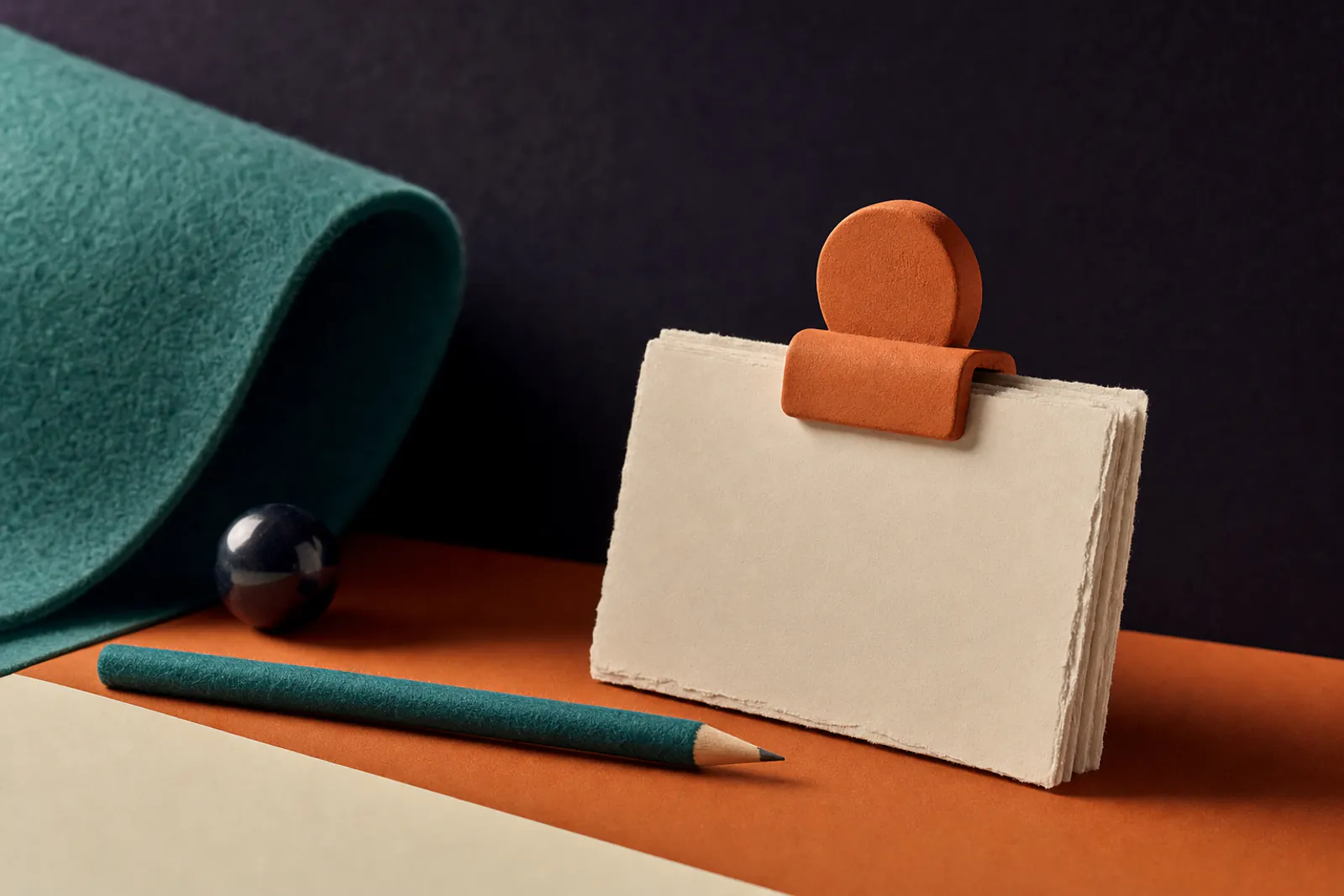 Illustration: a stack of cream paper cards with torn edges held by an orange clay clip, a teal felt pencil resting beside them.