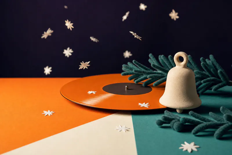 Illustration: a small cream clay bell and teal felt pine branches around an orange vinyl record, paper snowflakes falling.