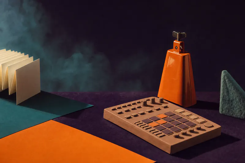 Illustration: a glossy tangerine cowbell and a clay drum machine on a dark violet field, faint teal haze like tire smoke.